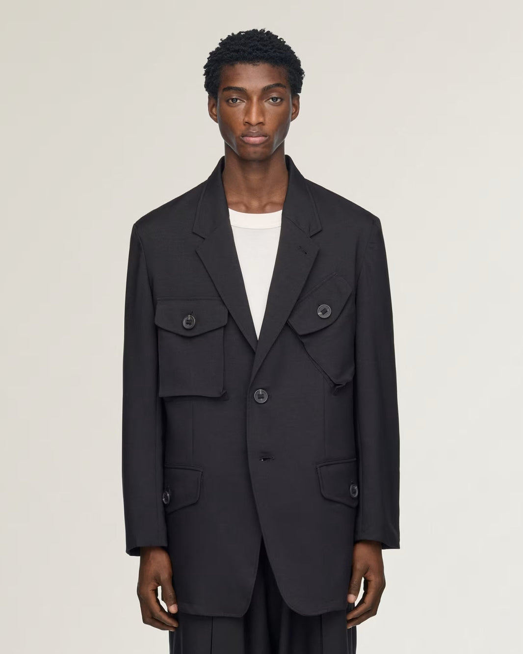 A model wearing a black double-breasted blazer with flap pockets over a white crewneck shirt, photographed against a solid light gray background.