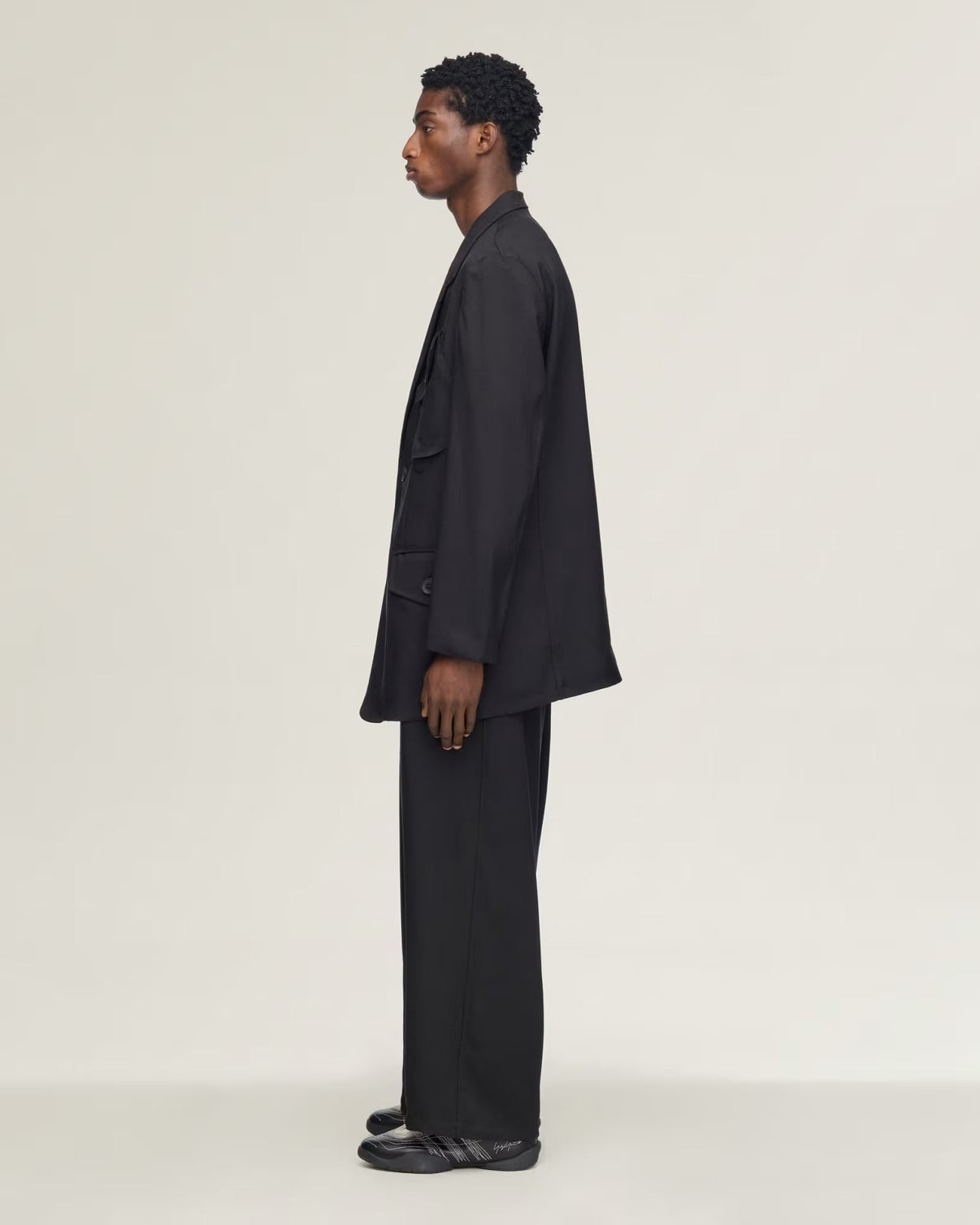 A model in a black oversized blazer and matching trousers, shown in a side profile against a plain light gray studio background.