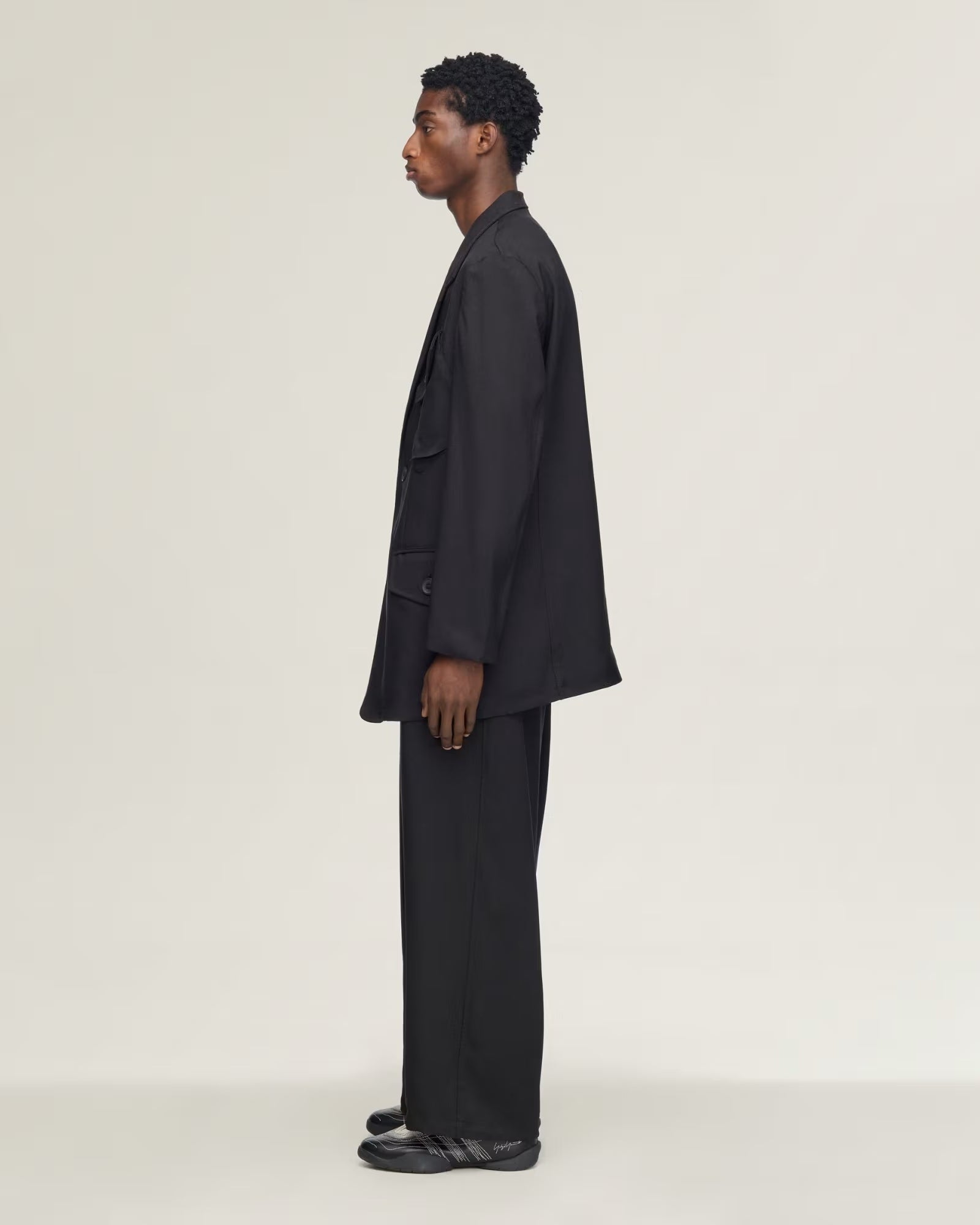 A model in a black oversized blazer and matching trousers, shown in a side profile against a plain light gray studio background.