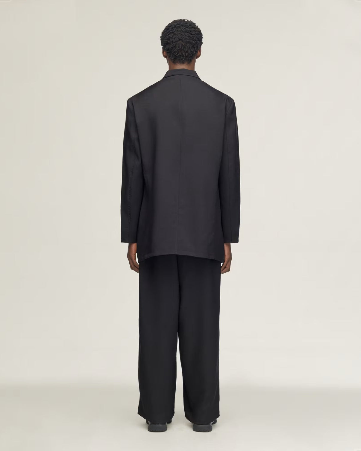 A model viewed from the back wearing a black long-sleeve blazer and matching wide-leg trousers against a plain light gray studio background.