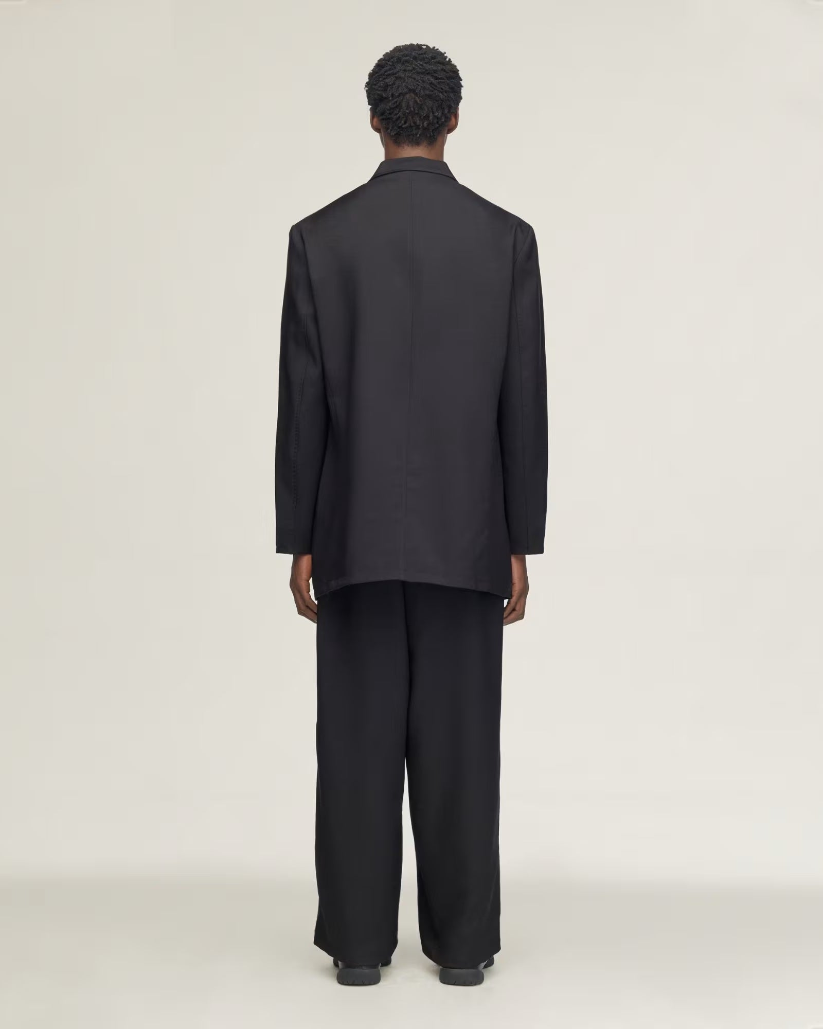 A model viewed from the back wearing a black long-sleeve blazer and matching wide-leg trousers against a plain light gray studio background.
