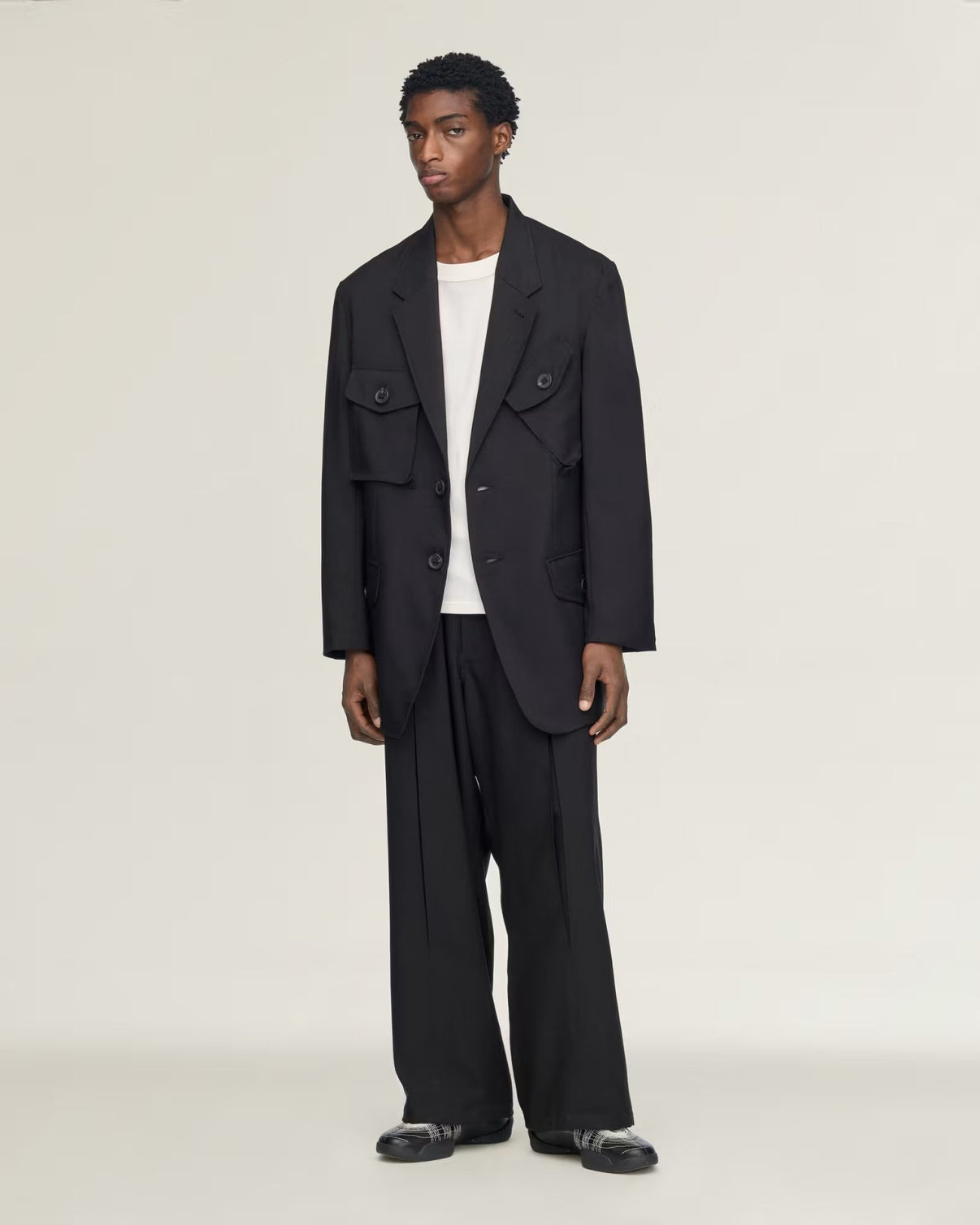 A model stands against a plain light gray studio background wearing a black double-breasted utility-style blazer with flap chest pockets, matching black wide-leg trousers, a white crew-neck undershirt, and black loafers.