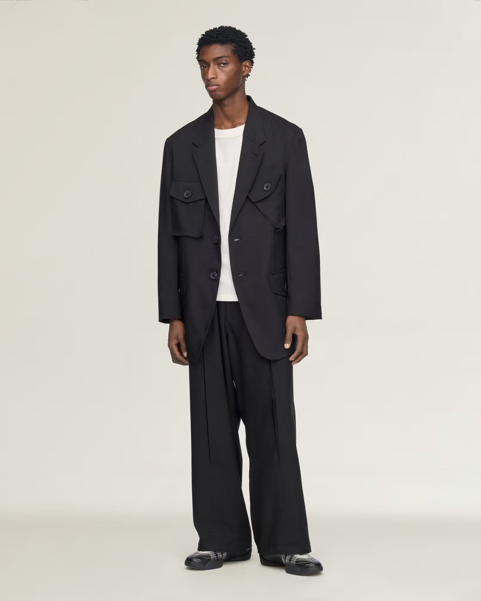 A model stands against a plain light gray studio background wearing a black double-breasted utility-style blazer with flap chest pockets, matching black wide-leg trousers, a white crew-neck undershirt, and black loafers.