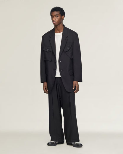 A model stands against a plain light gray studio background wearing a black double-breasted utility-style blazer with flap chest pockets, matching black wide-leg trousers, a white crew-neck undershirt, and black loafers.