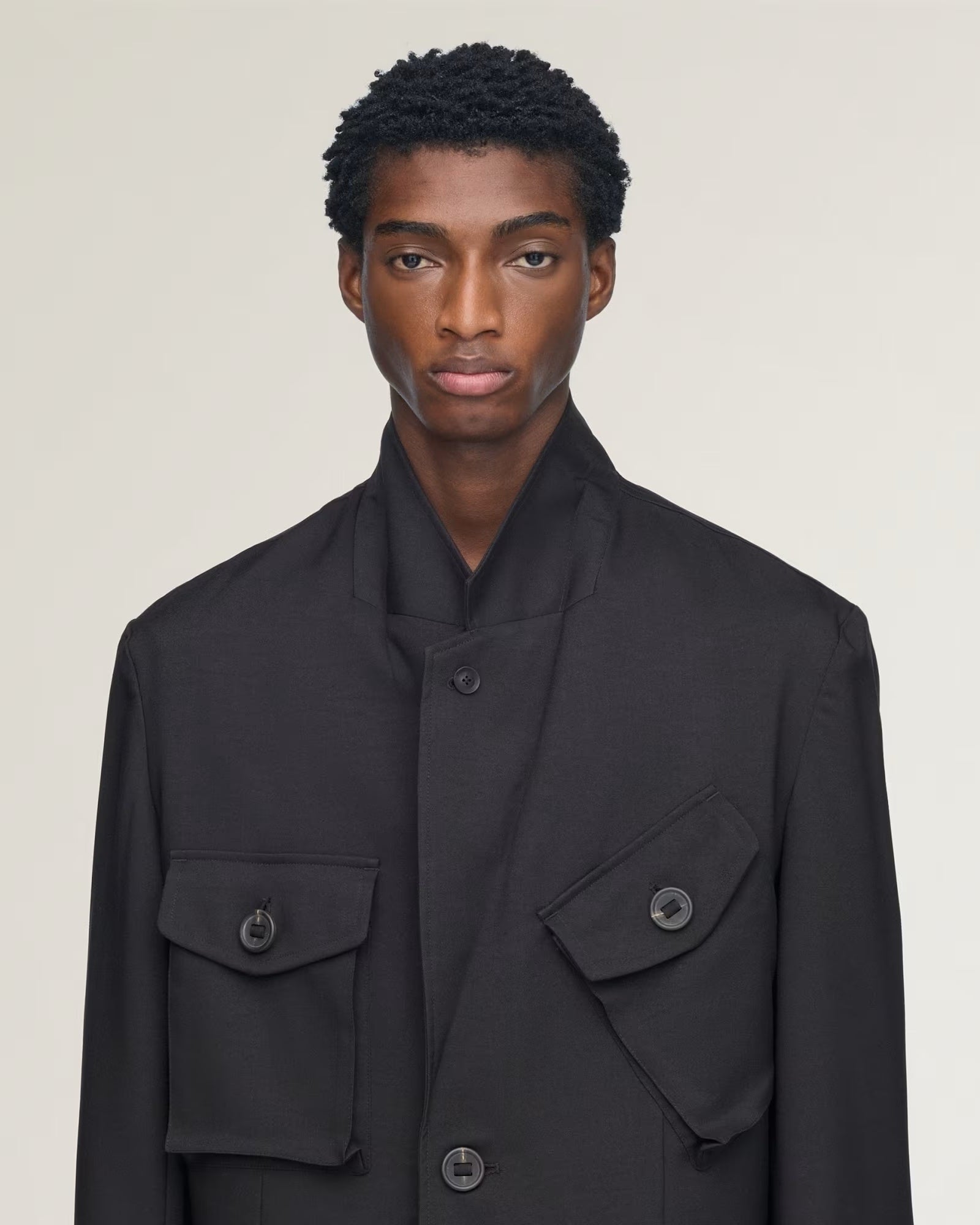 A model wearing a black structured jacket with a high stand collar, button front, and two large flap pockets on a light beige studio background.