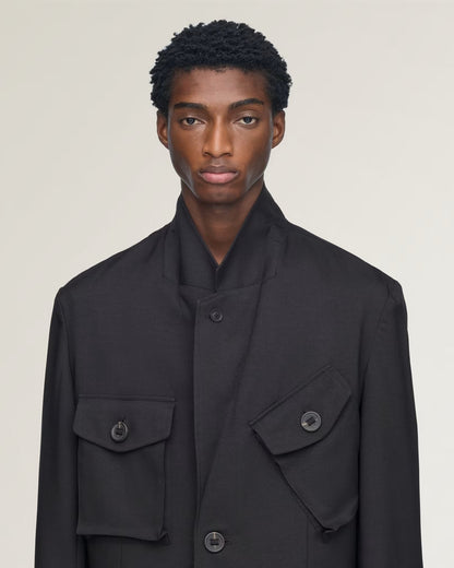 A model wearing a black structured jacket with a high stand collar, button front, and two large flap pockets on a light beige studio background.