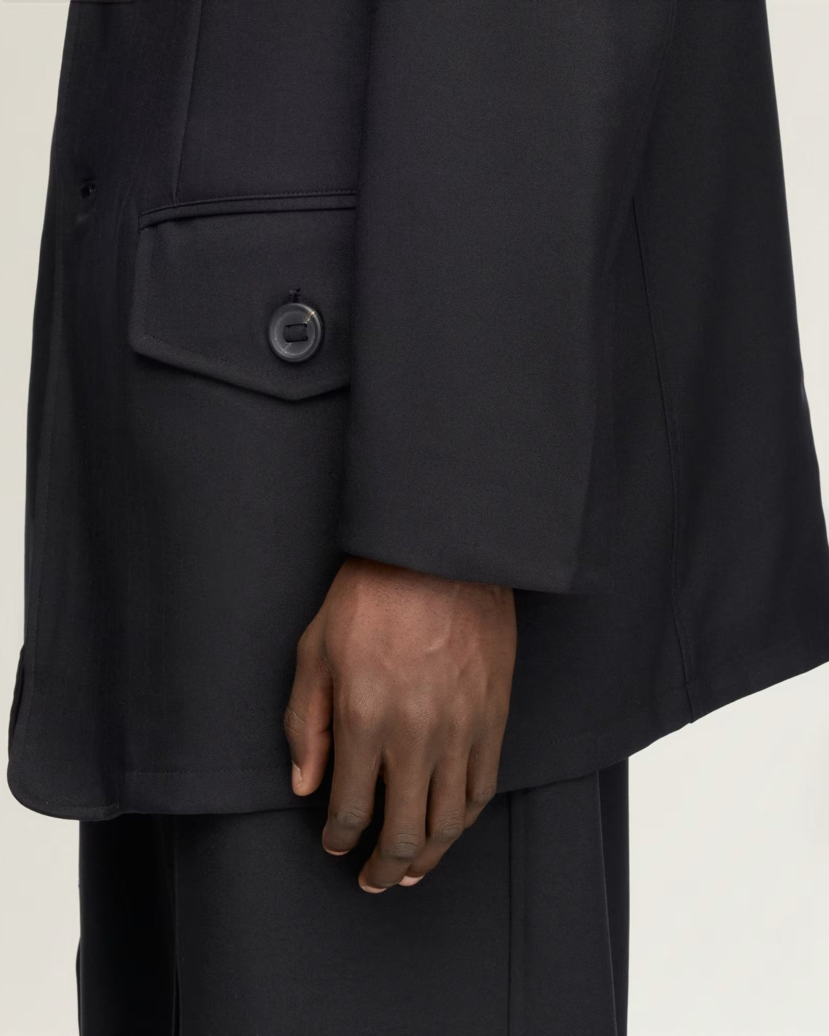 Close-up of a person wearing an oversized black blazer with a single-breasted flap pocket and matching black trousers, against a plain light background.