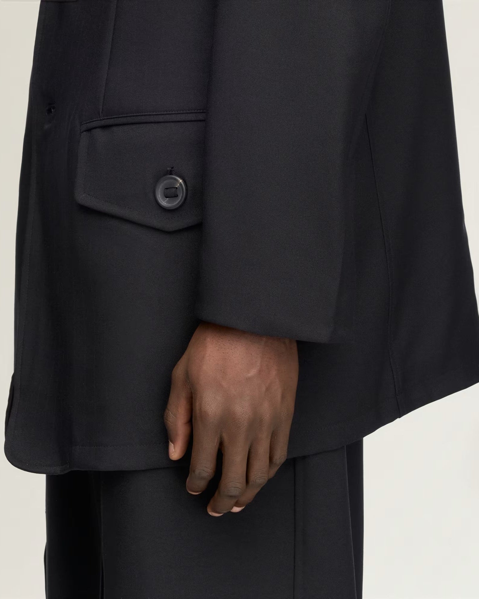 Close-up of a person wearing an oversized black blazer with a single-breasted flap pocket and matching black trousers, against a plain light background.