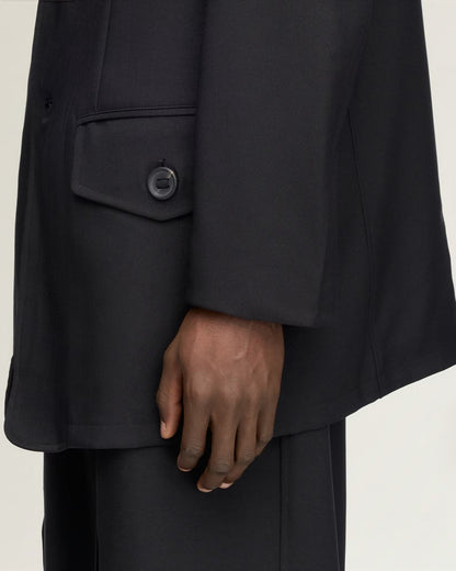 Close-up of a person wearing an oversized black blazer with a single-breasted flap pocket and matching black trousers, against a plain light background.