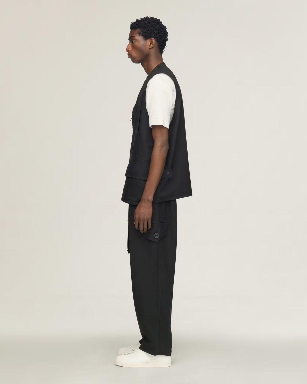 A man in profile wearing a black sleeveless vest over a white short-sleeve shirt, black trousers, and white sneakers, standing against a plain light gray studio background.