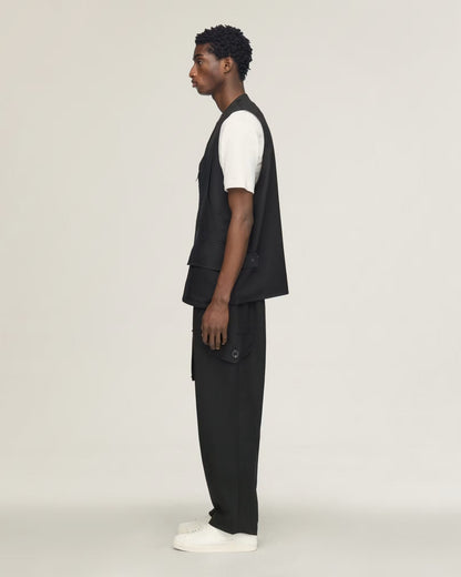 A man in profile wearing a black sleeveless vest over a white short-sleeve shirt, black trousers, and white sneakers, standing against a plain light gray studio background.