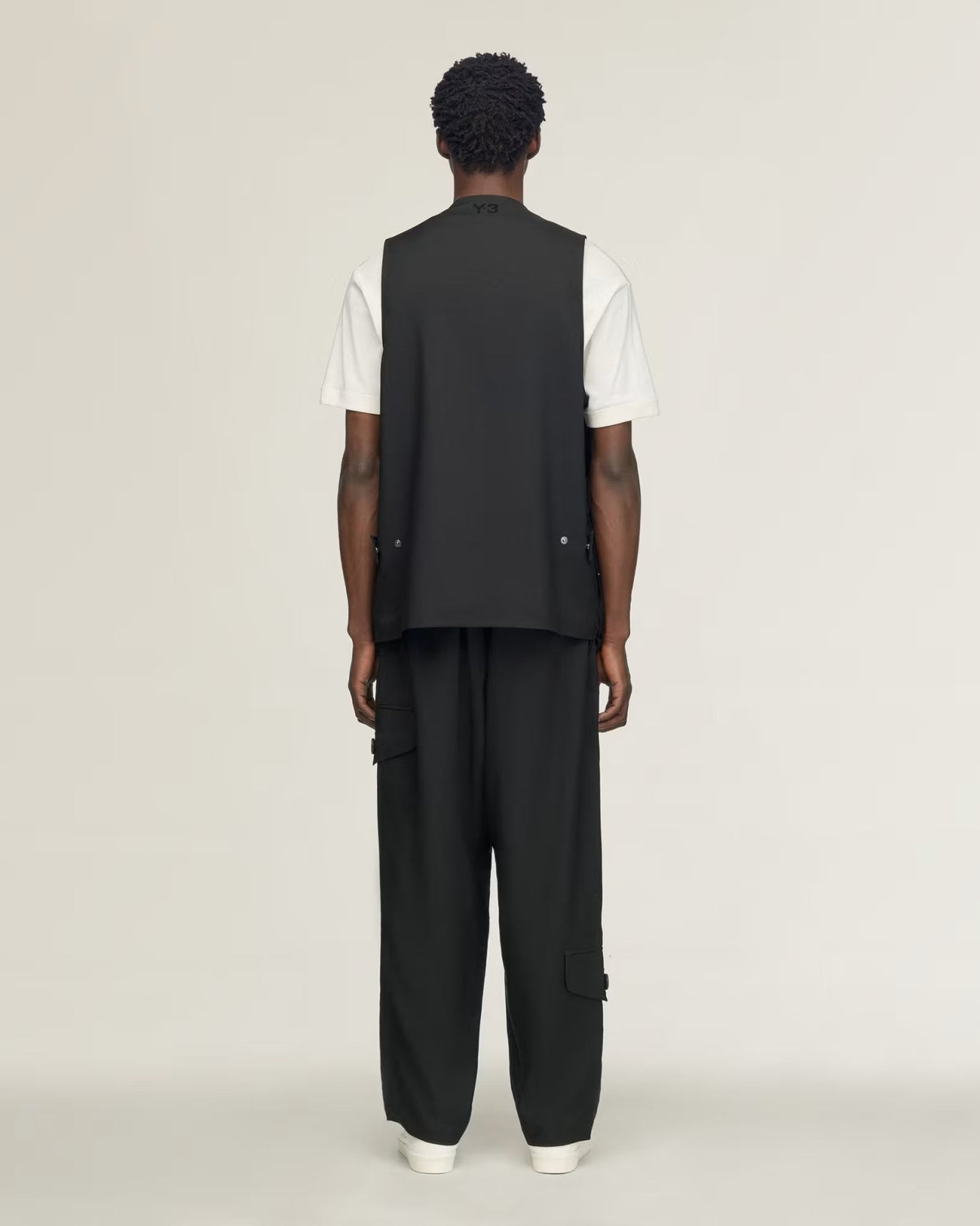 A model shown from the back wearing a black sleeveless vest over a white t-shirt and black wide-leg cargo pants, set against a plain light gray studio background.